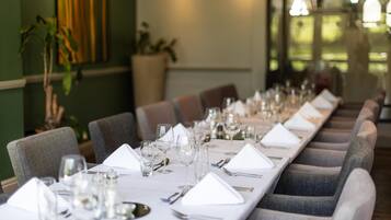 A long dining table set with white tablecloths, silverware, and wine glasses, flanked by upholstered chairs.