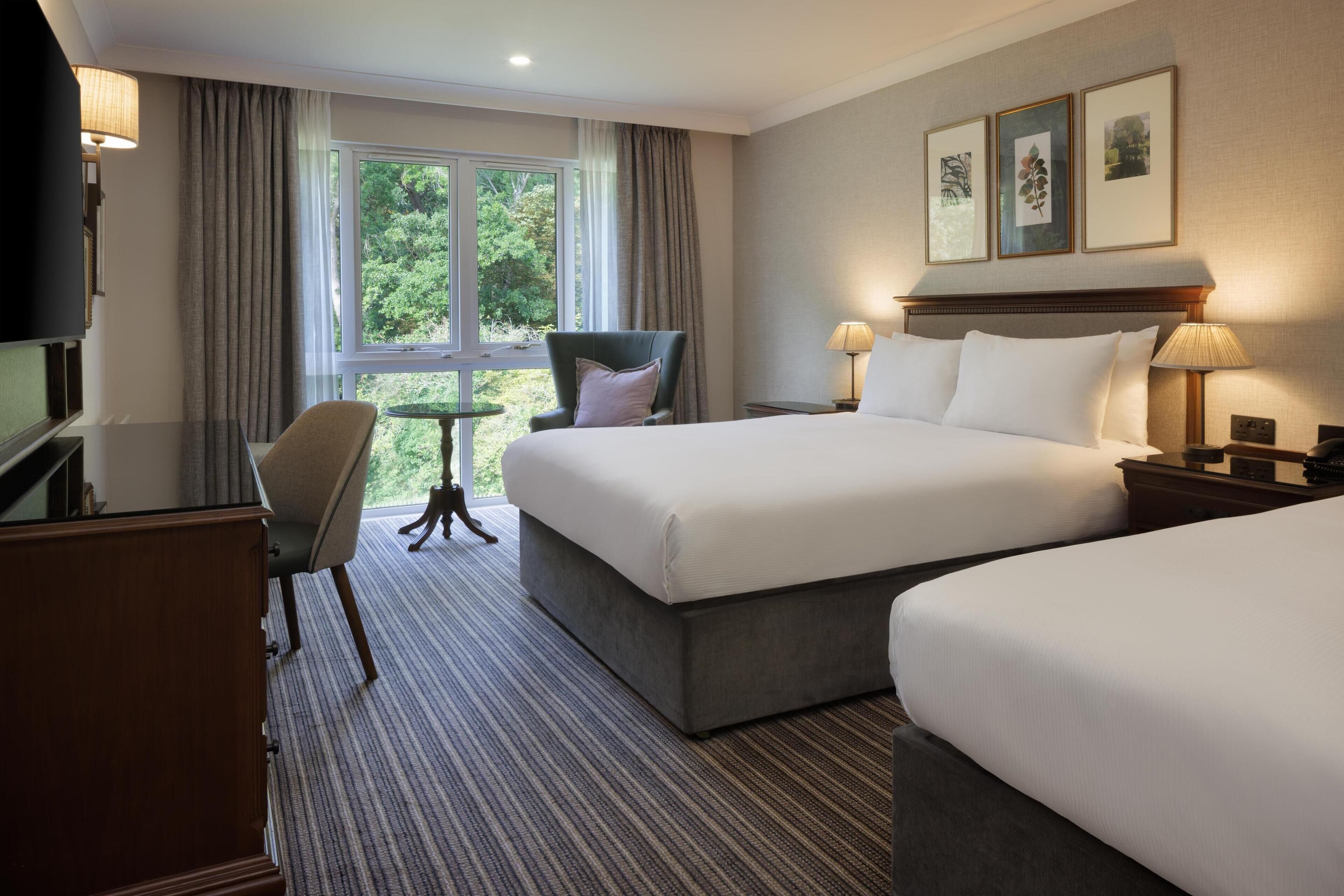 A hotel room with two beds, a desk, a chair, and a window with a view of greenery.