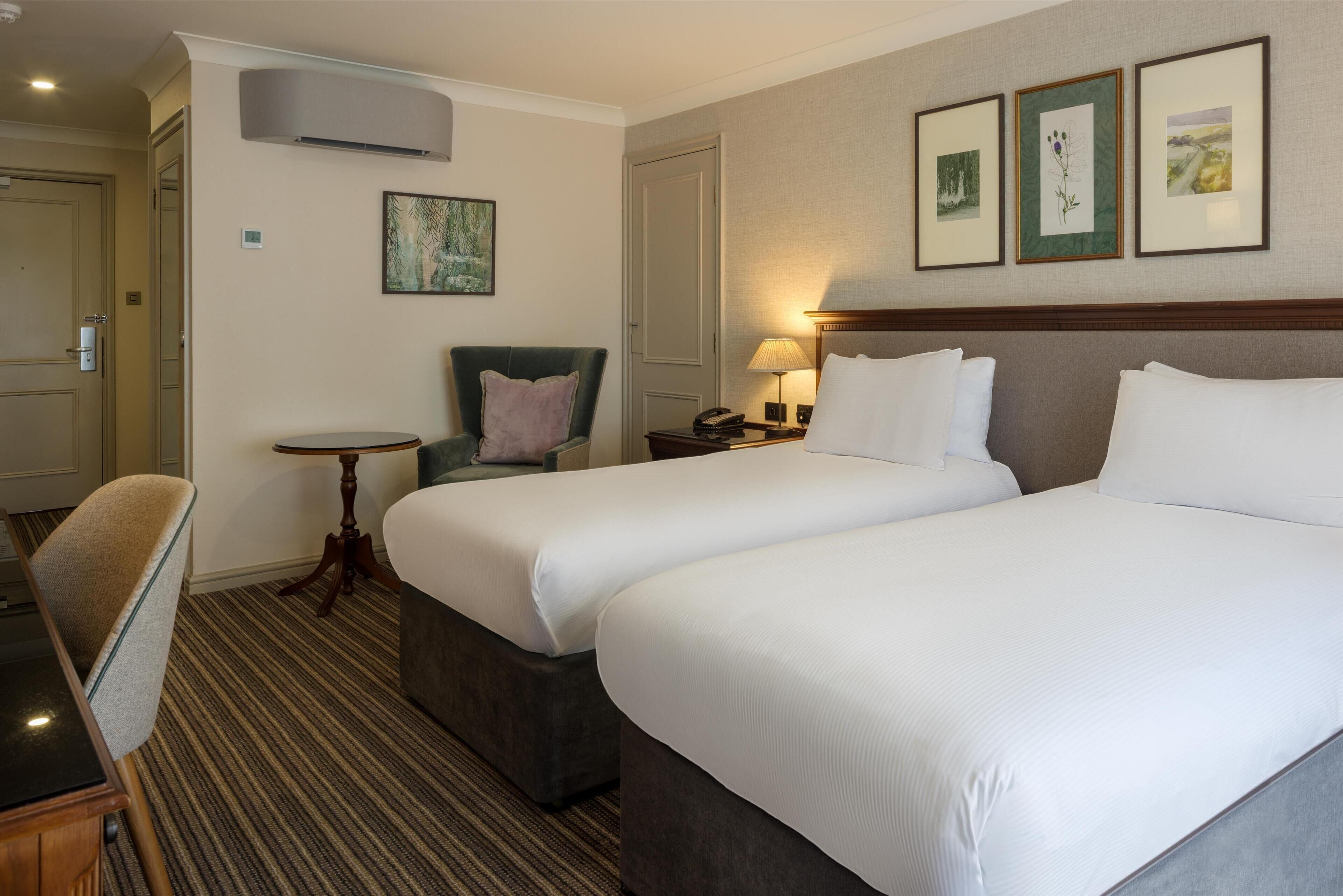 Habitación de hotel con dos camas, una silla, una mesita pequeña y cuadros en la pared.