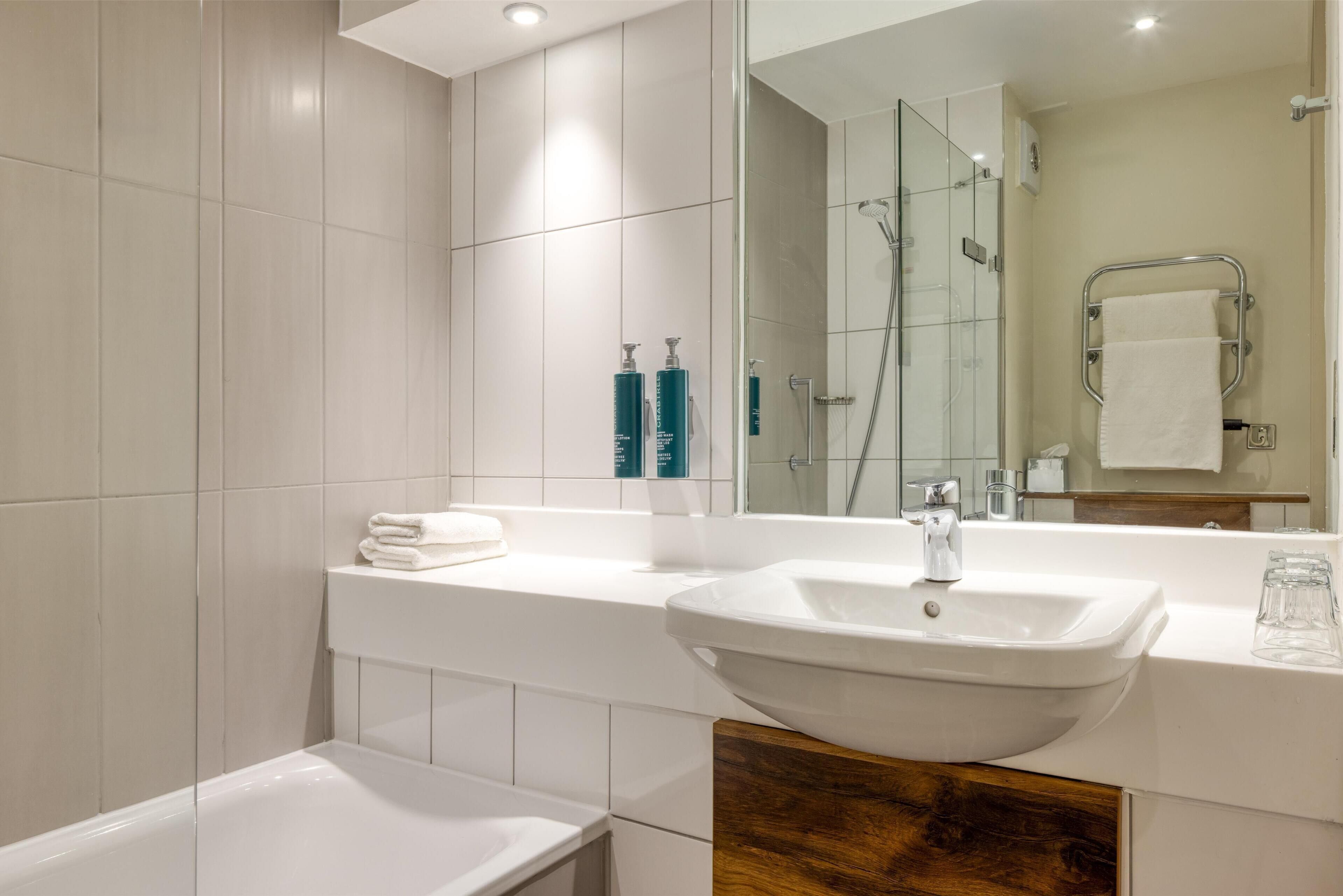 A modern bathroom with a large mirror, white tiles, a white sink, and a glass-enclosed shower.