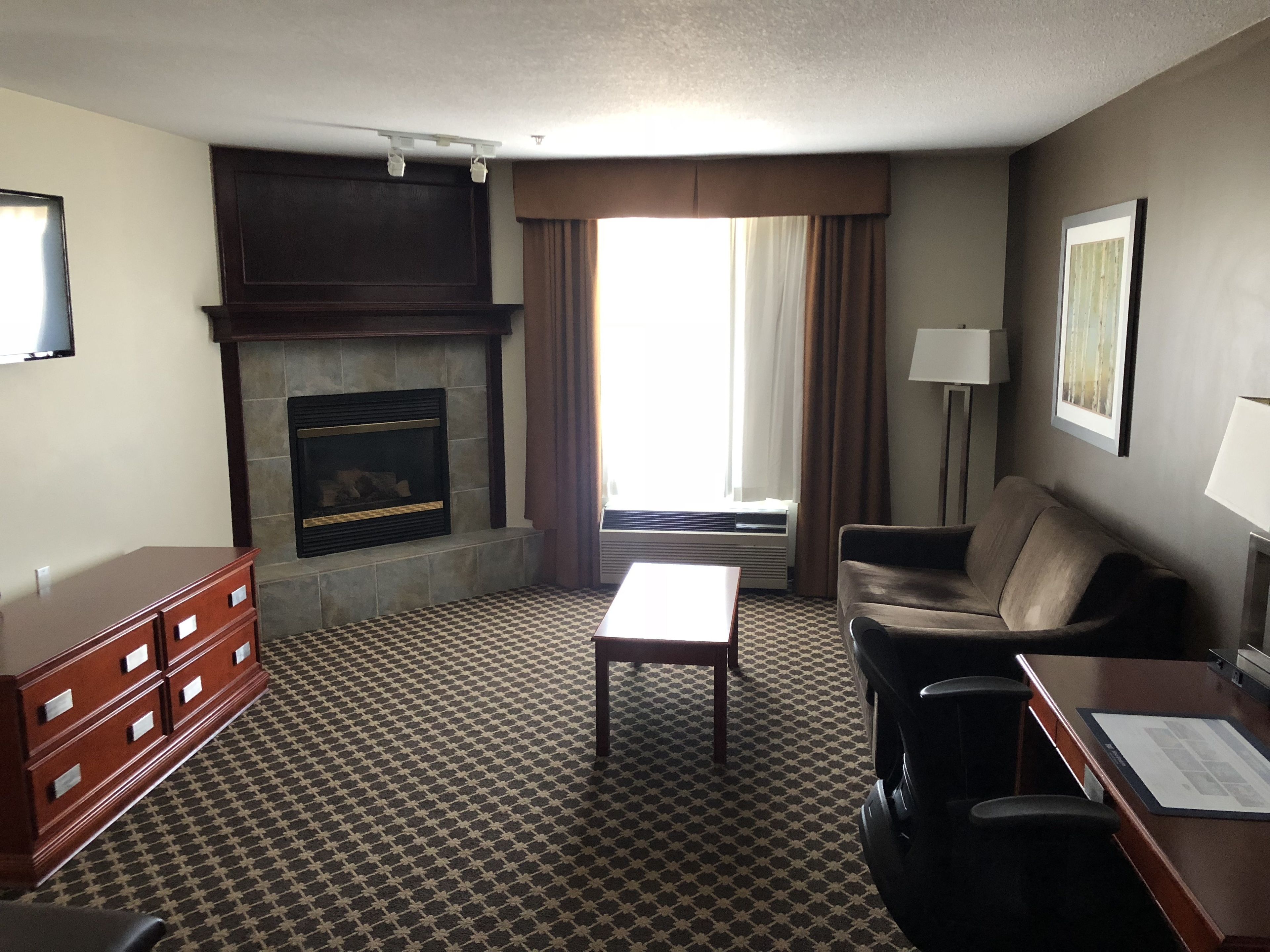 A hotel room with a fireplace, a sofa, a coffee table, a desk with a chair, and a patterned carpet.