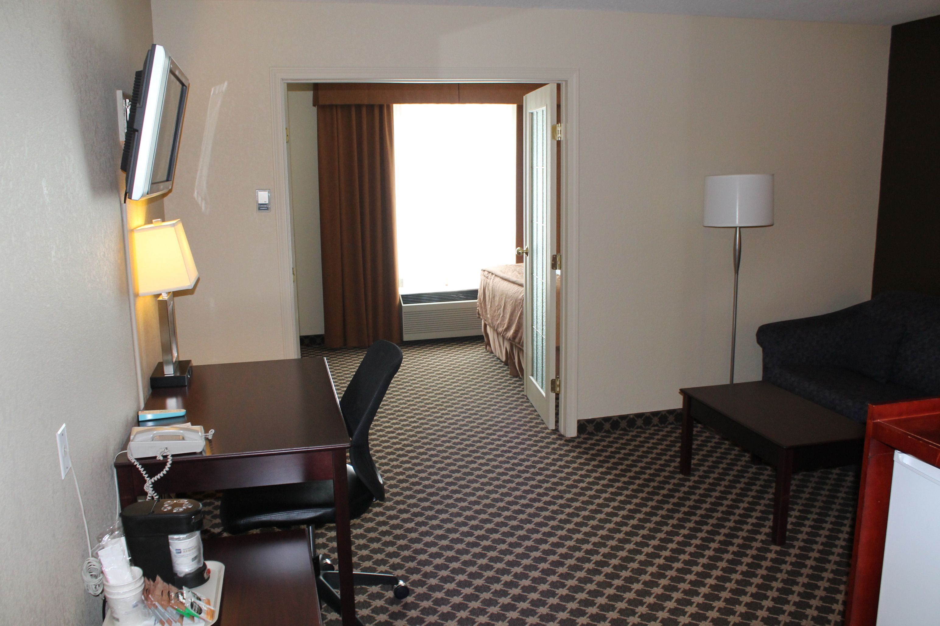 A hotel room with a desk, chair, lamp, and a couch. There is a window with curtains, a door leading to another room, and a patterned carpet.