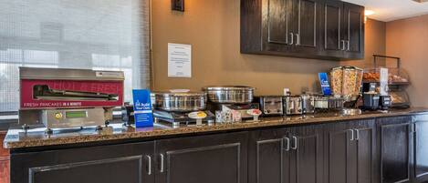 A hotel kitchen with a self-serve breakfast station, including a hot pancake machine and various food containers.