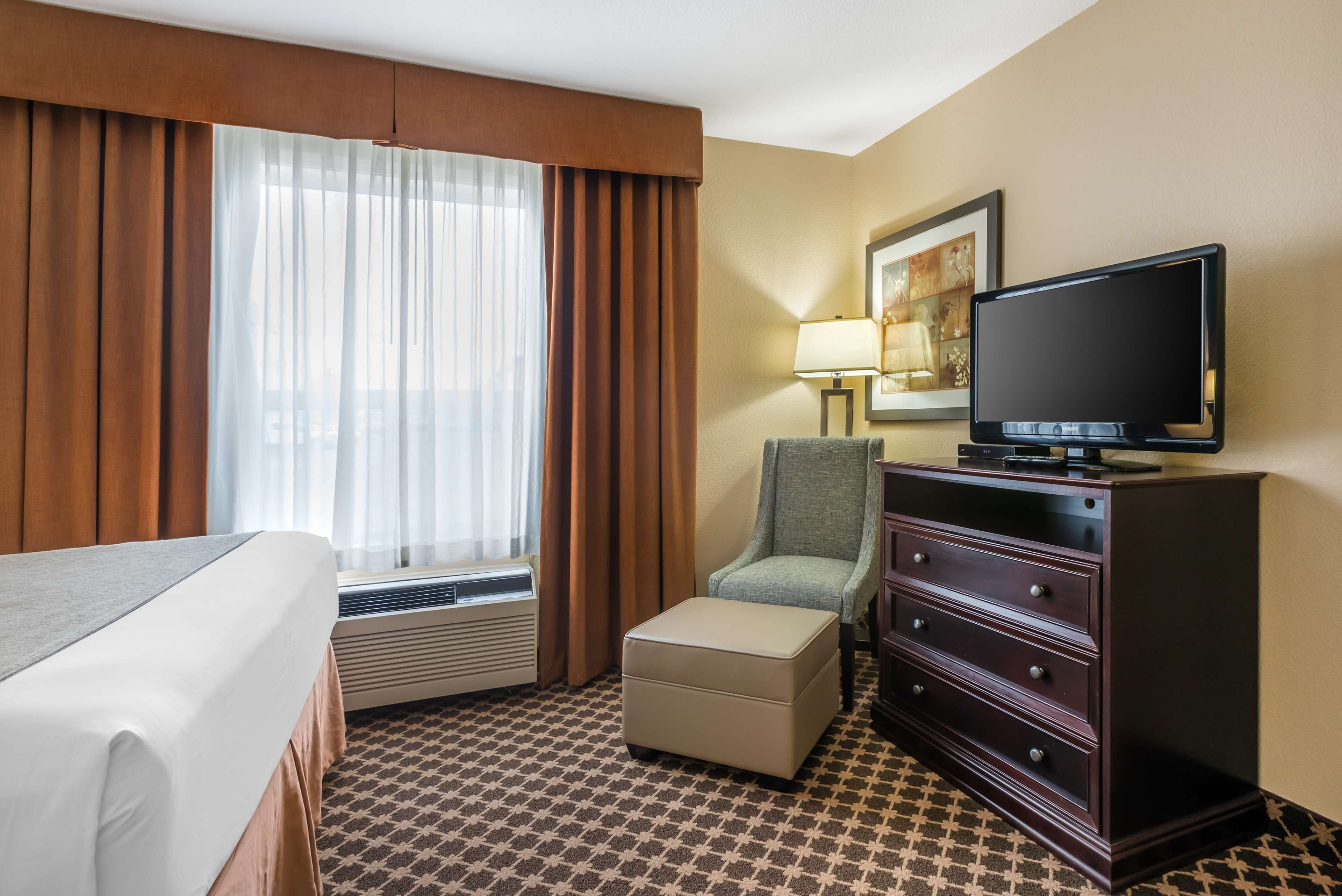 A hotel room with a bed, a television on a dresser, a chair, a lamp, and a painting on the wall.