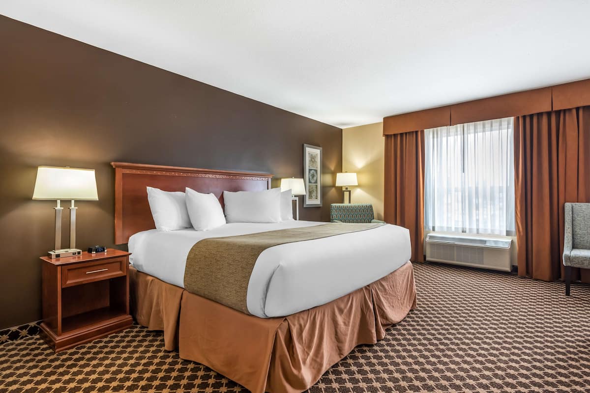 Ein Hotelzimmer mit einem großen Bett, zwei Nachttischen inklusive Lampen, einem Sessel, einem Fenster mit Vorhängen und einer Klimaanlage.