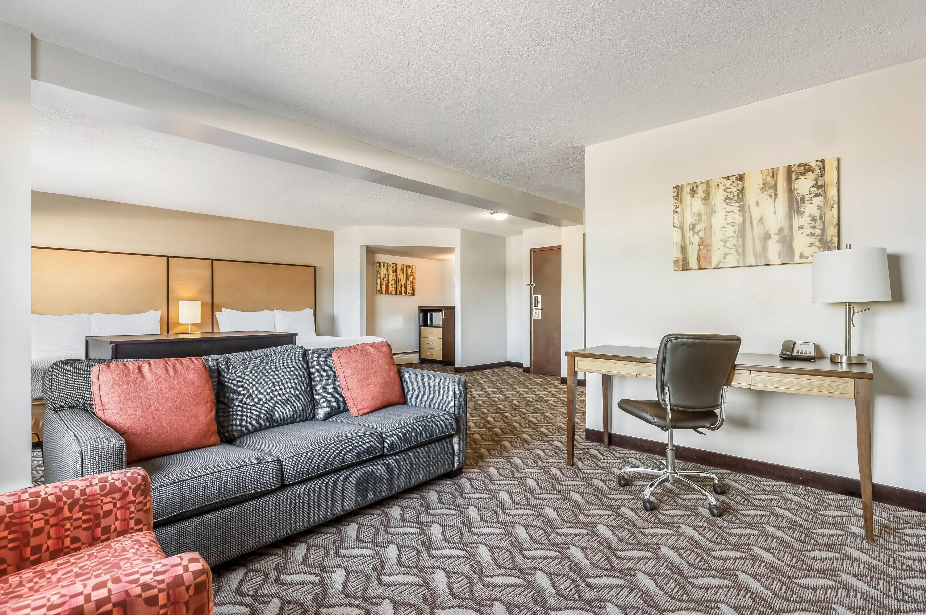 A hotel room with a grey sofa, a patterned rug, a desk with a chair, and a painting on the wall.
