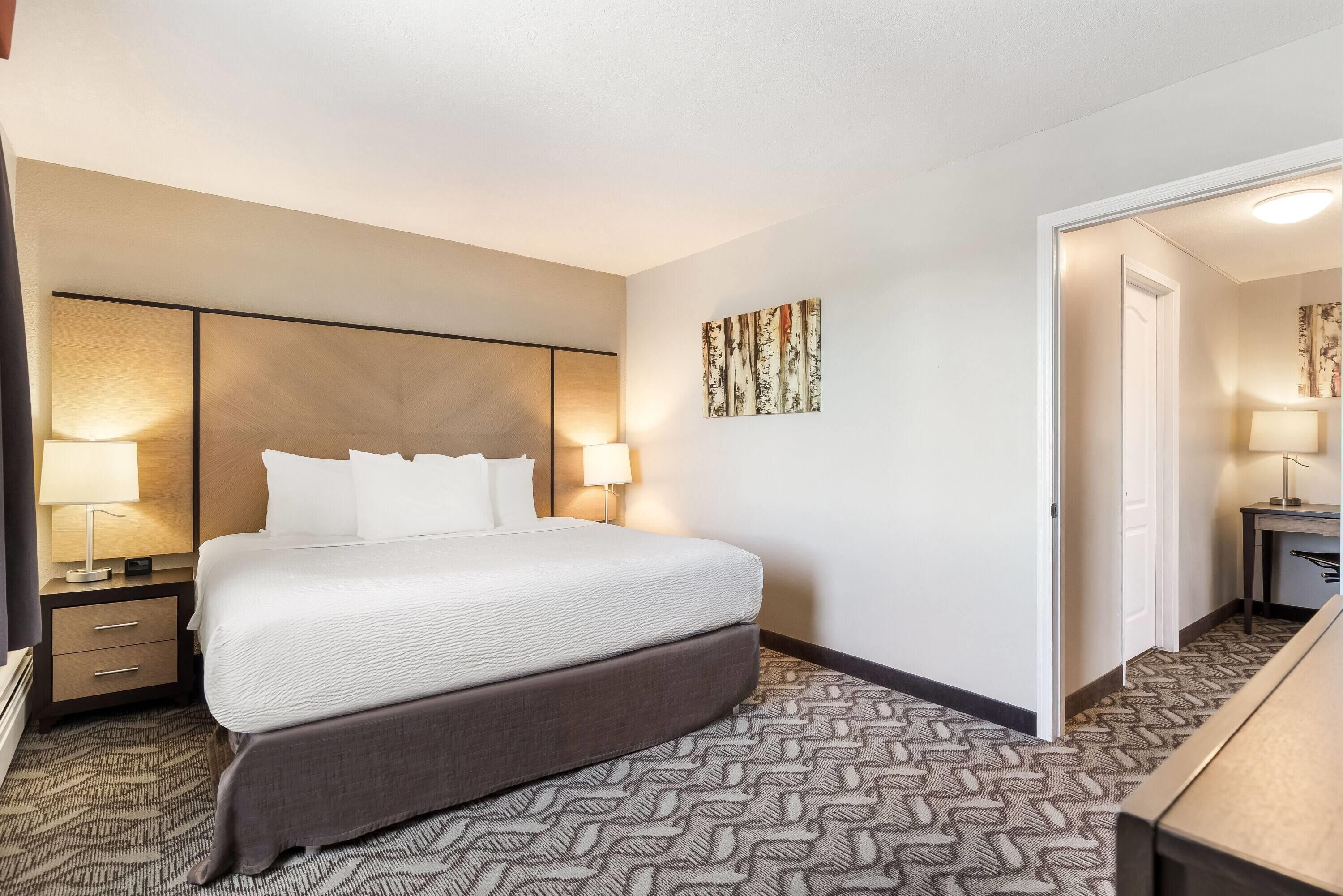 Una habitación de hotel con una cama grande, una mesita de noche con lámpara, un cabecero, una alfombra estampada y una puerta que da a otra habitación.