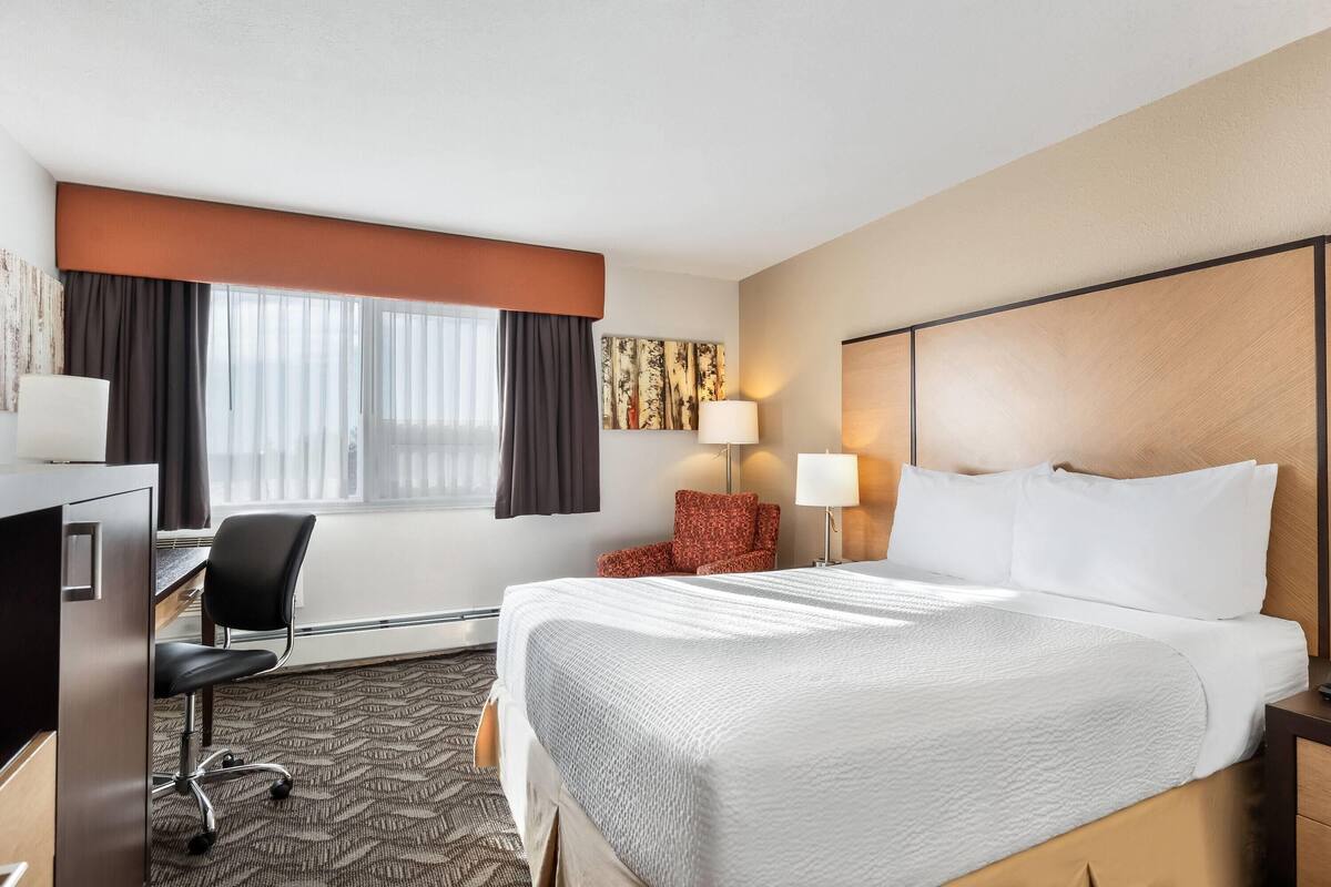 A hotel room with a large bed, a desk with a chair, a lamp, and a window with curtains.