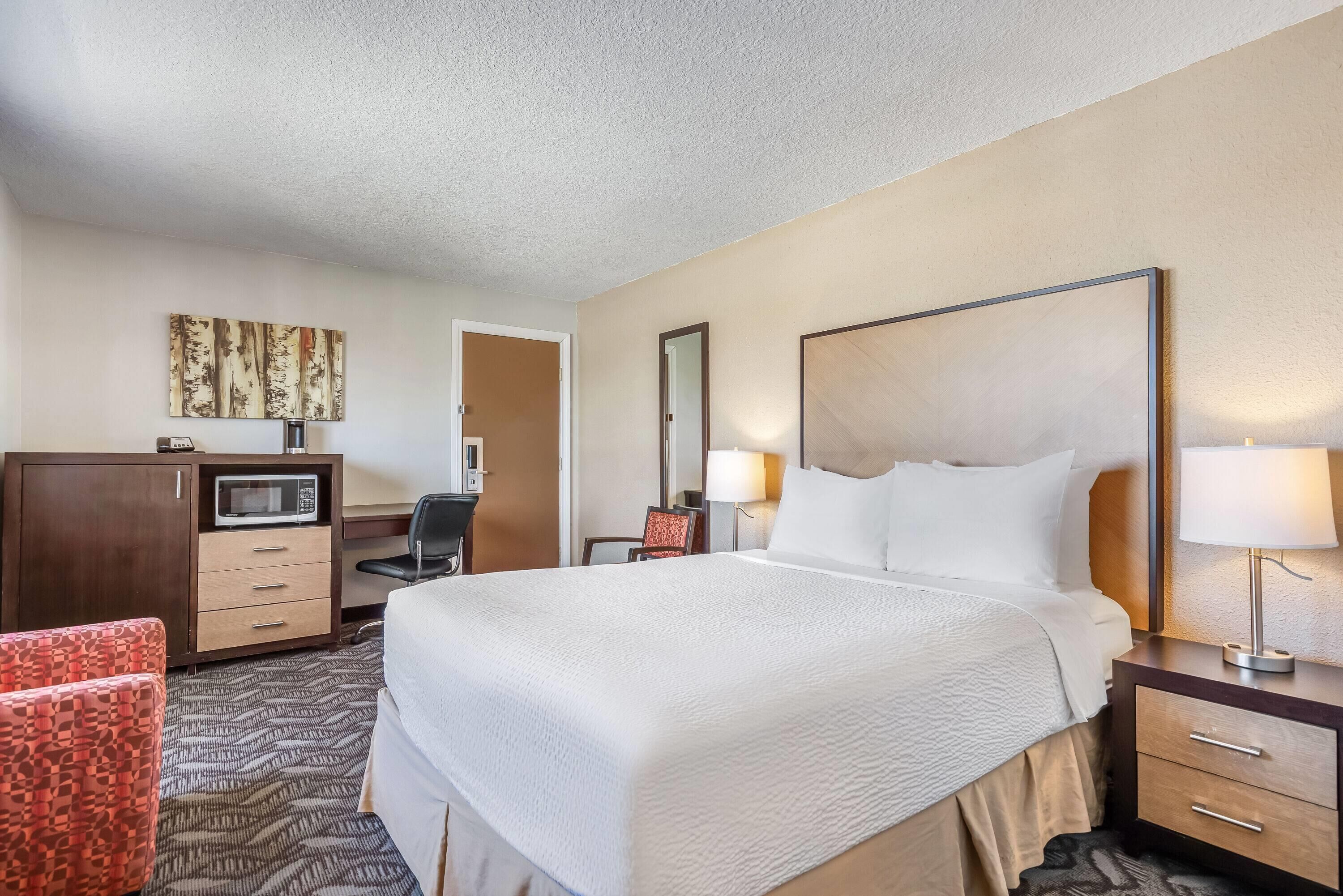 A hotel room with a large bed, a desk with a microwave, a chair, and a desk lamp.