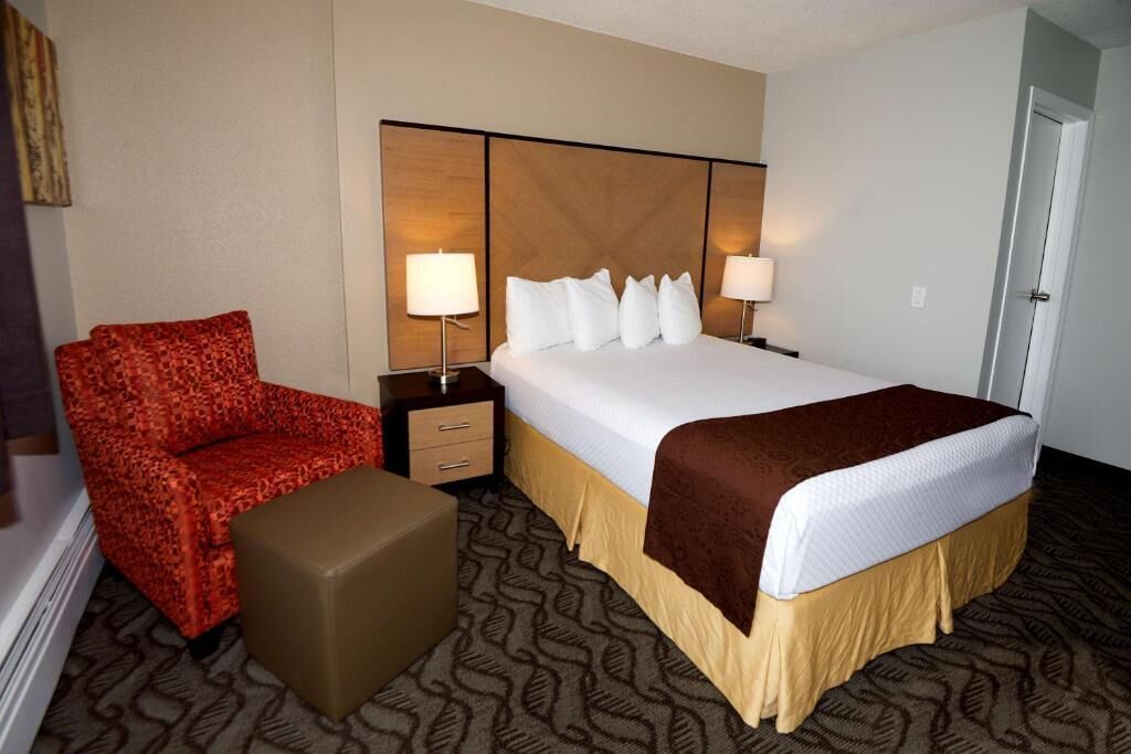 A hotel room with a bed, two bedside tables with lamps, a red patterned sofa, a brown ottoman, and a patterned carpet.