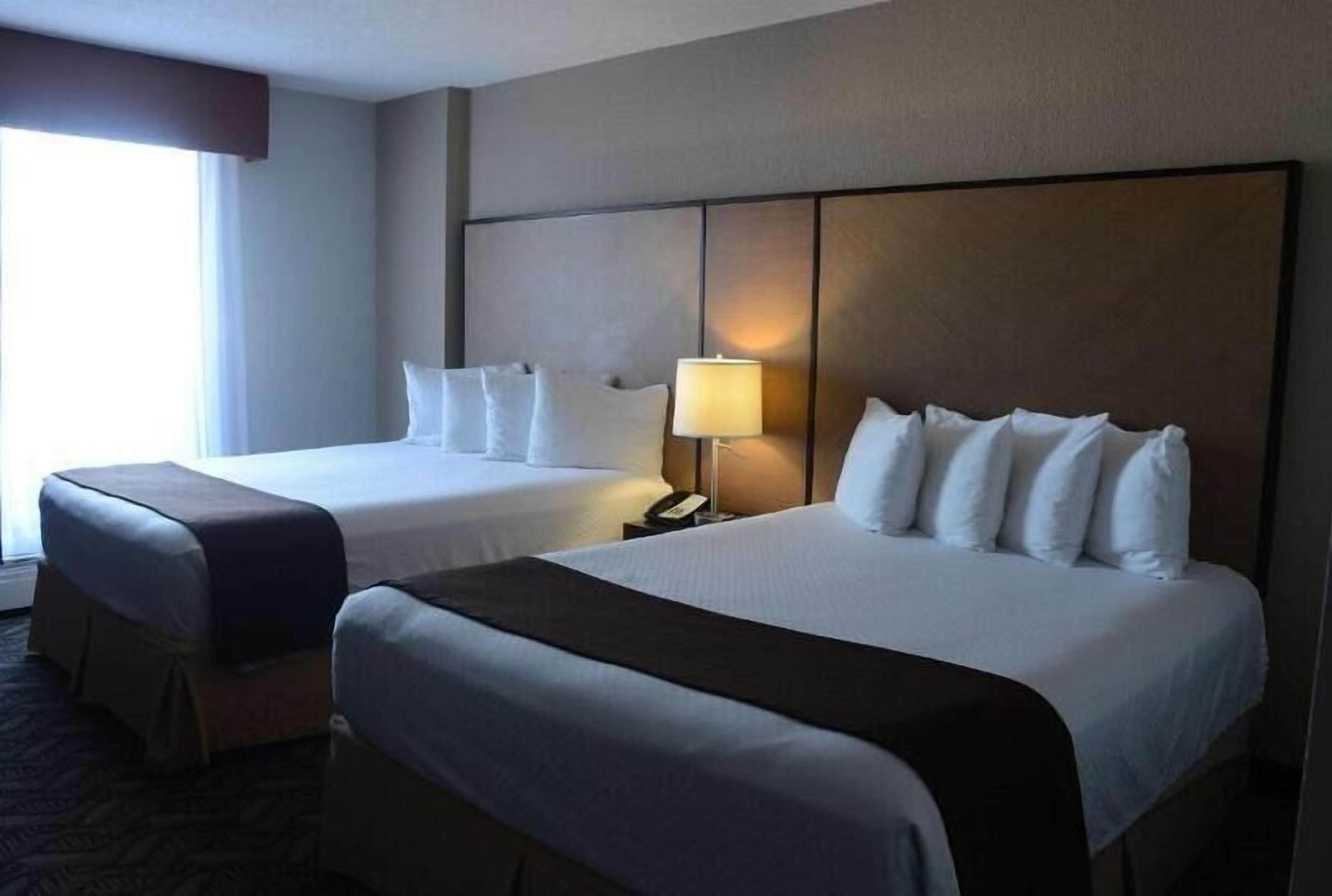 Habitación de hotel con dos camas, una mesita de noche con lámpara y ventana que deja entrar luz natural.