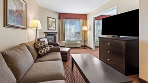 A hotel room with a sofa, a TV on a dresser, a fireplace, and a large window with curtains. - Best Western Of Olds (Olds)