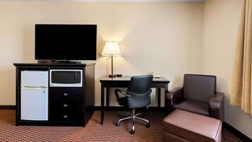 Quarto de hotel com televisão de tela plana, geladeira, micro-ondas, escrivaninha com luminária, cadeira de escritório preta, poltrona de couro marrom e um banquinho marrom.