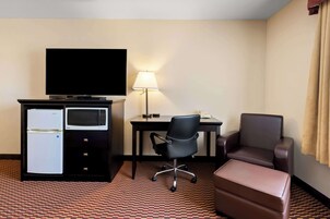 Quarto de hotel com televisão de tela plana, geladeira, micro-ondas, escrivaninha com luminária, cadeira de escritório preta, poltrona de couro marrom e um banquinho marrom.