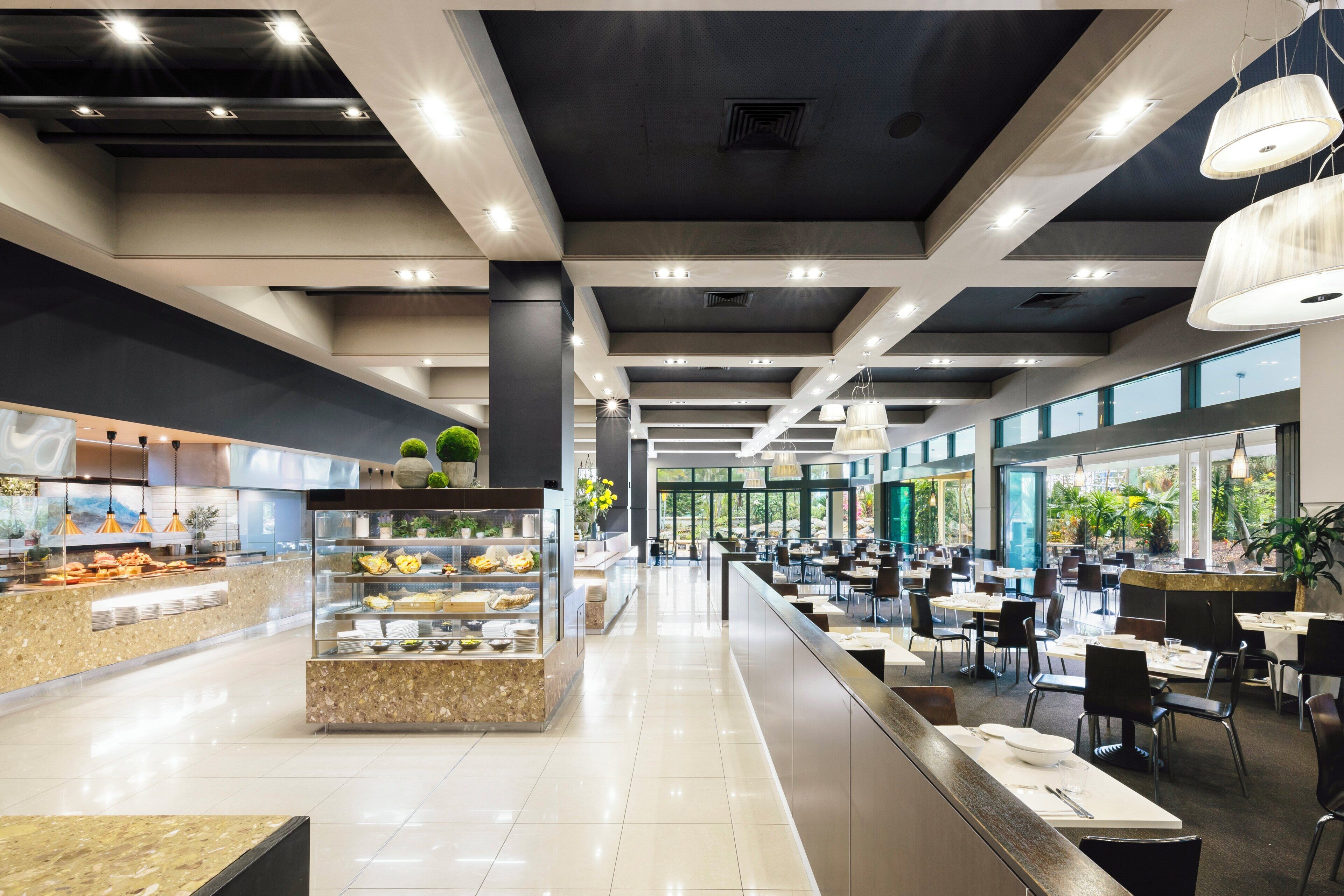 Una cafetería moderna con un área de servicio central, diferentes opciones de asientos y amplios ventanales que permiten la entrada de luz natural.