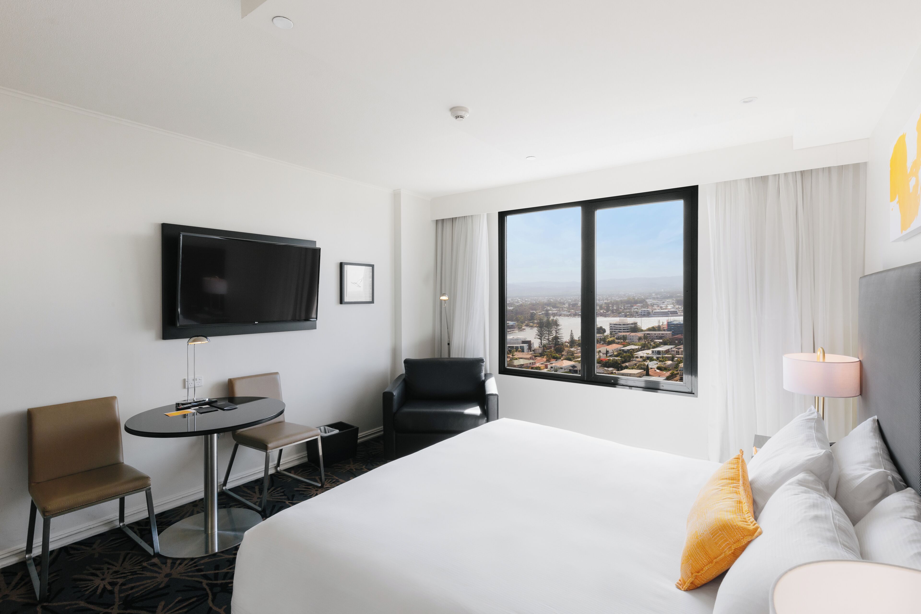 A hotel room with a bed, a TV, a small table, two chairs, and a large window with a city view.