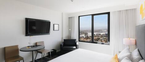 A hotel room with a bed, a TV, a small table, two chairs, and a large window with a city view.