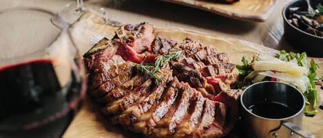 A wooden cutting board with sliced steak, a glass of red wine, and a small metal cup.