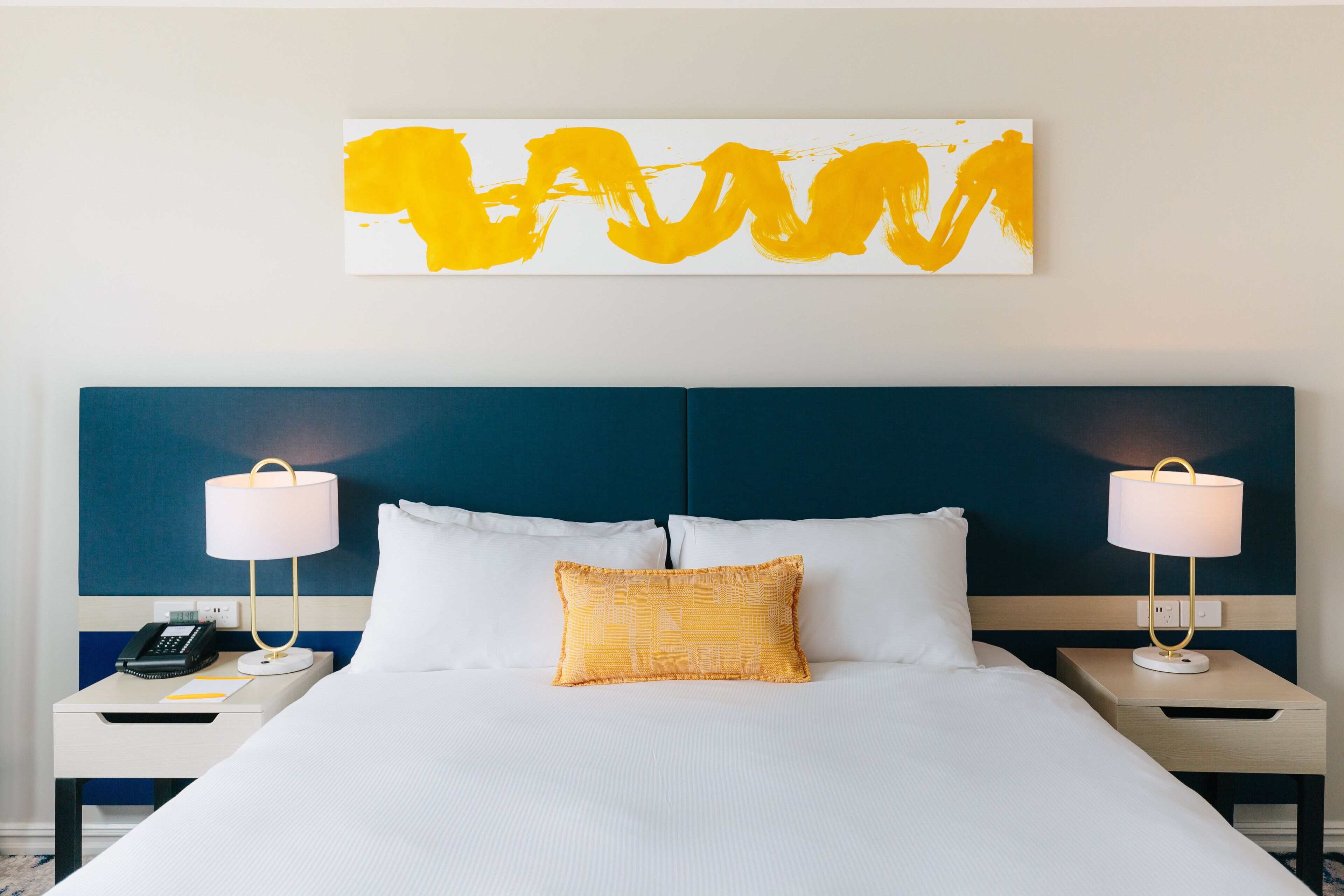 Een opgemaakt bed met witte lakens, twee bedlampjes, een telefoon en een geel kussen.