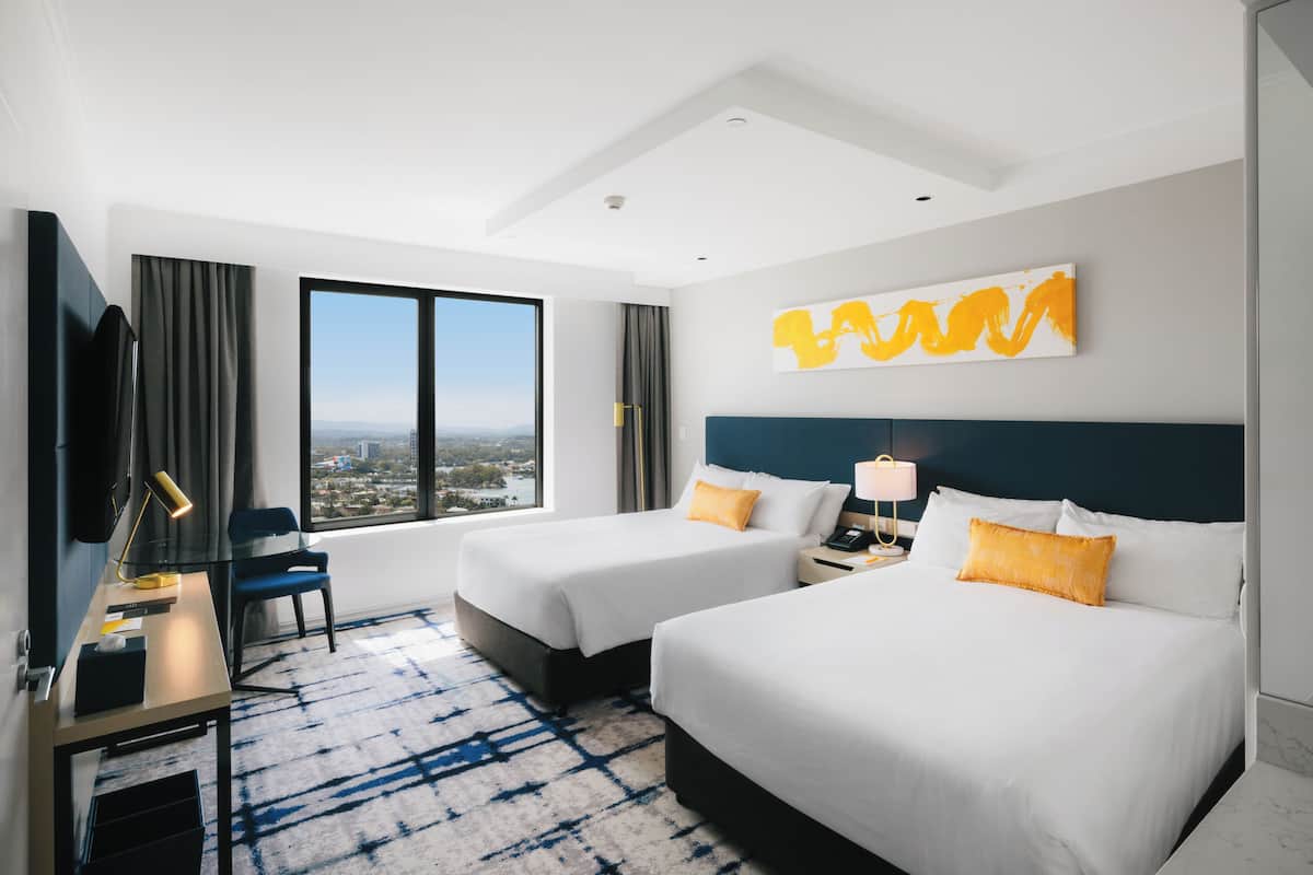 A hotel room with two beds, a desk, a chair, a TV, and a large window with a view.