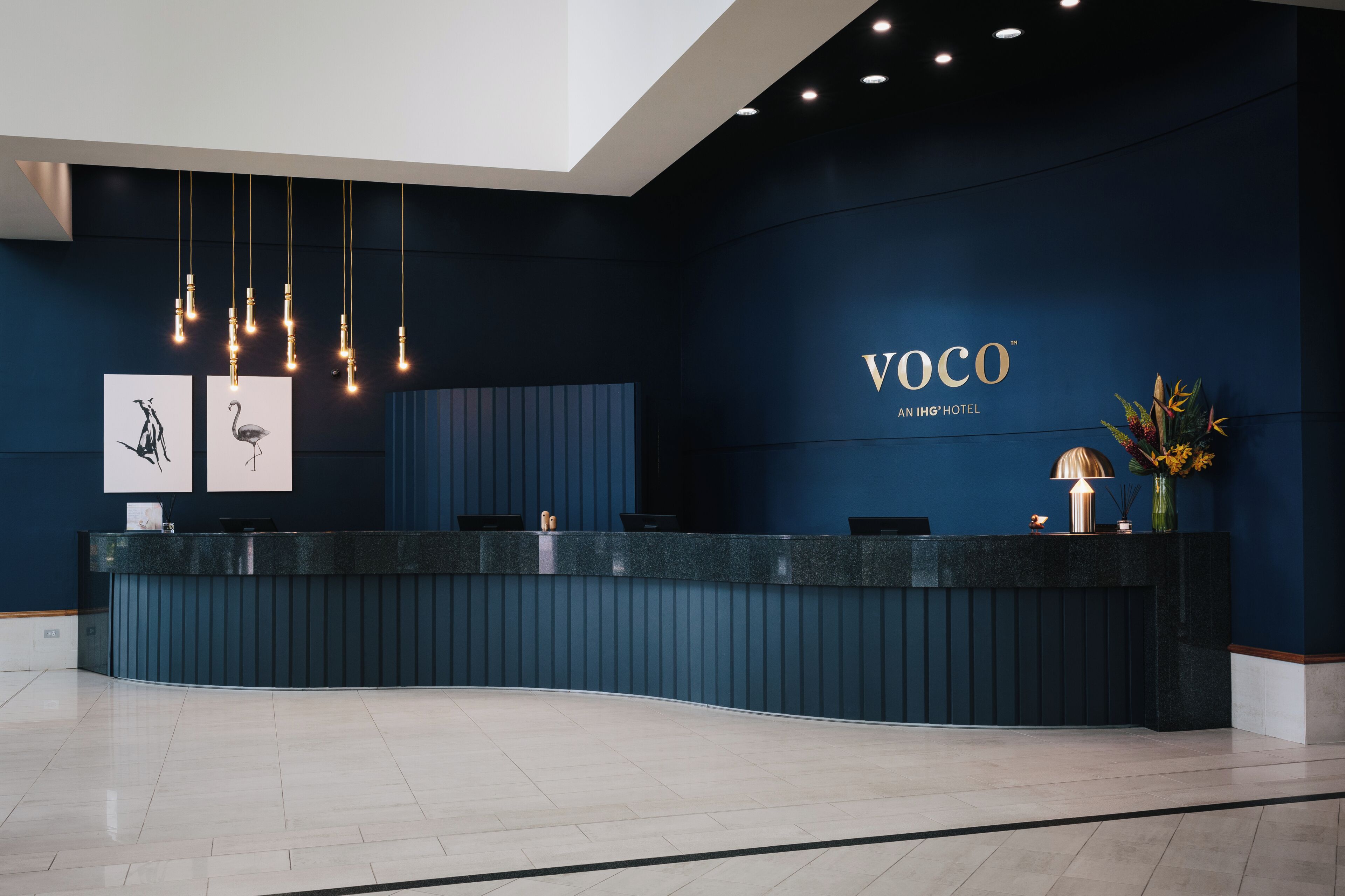 A reception desk with a dark blue wall, a sign reading 'VOCO AN IHG HOTEL', and a vase with flowers.