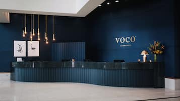 A reception desk with a dark blue wall, a sign reading 'VOCO AN IHG HOTEL', and a vase with flowers.