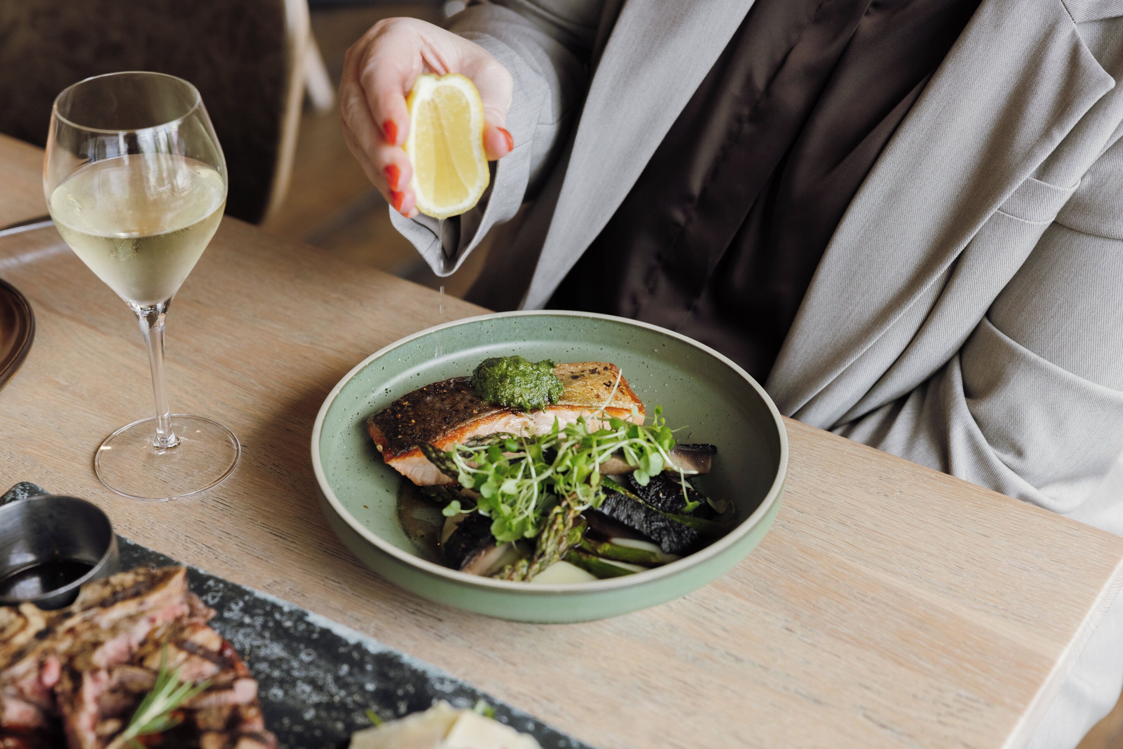 A dining table with a bowl of food, a glass of white wine, and a person holding a lemon.