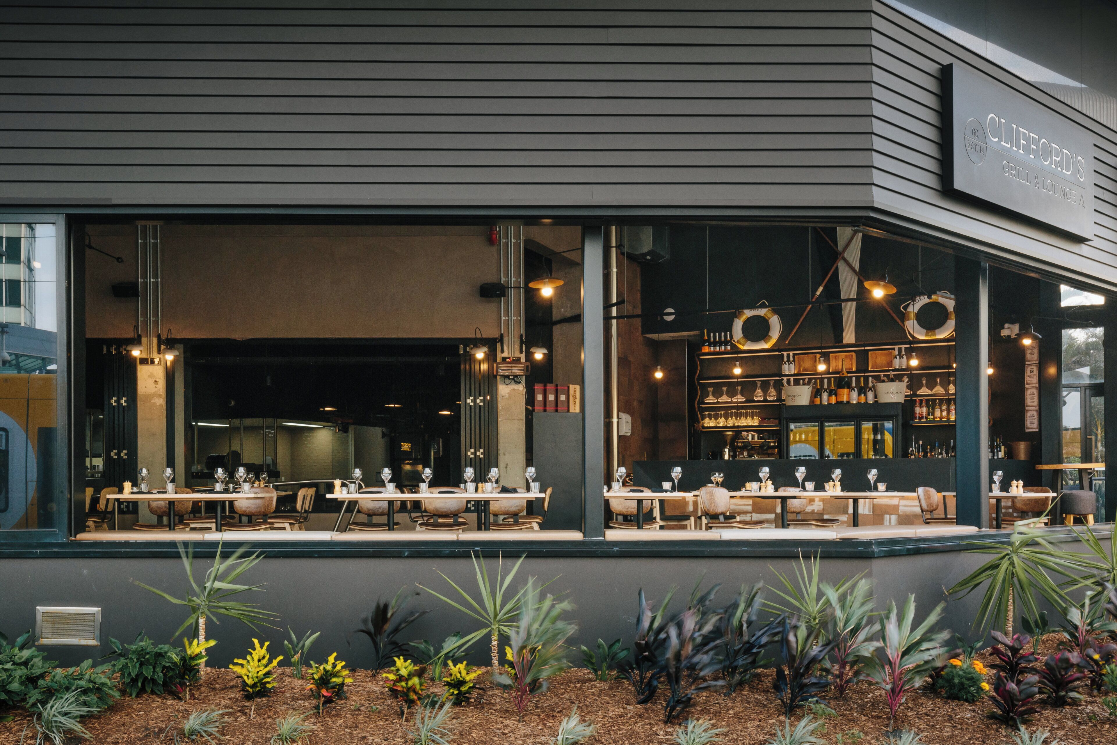 A restaurant with outdoor seating, a bar area, and a variety of plants in the foreground.