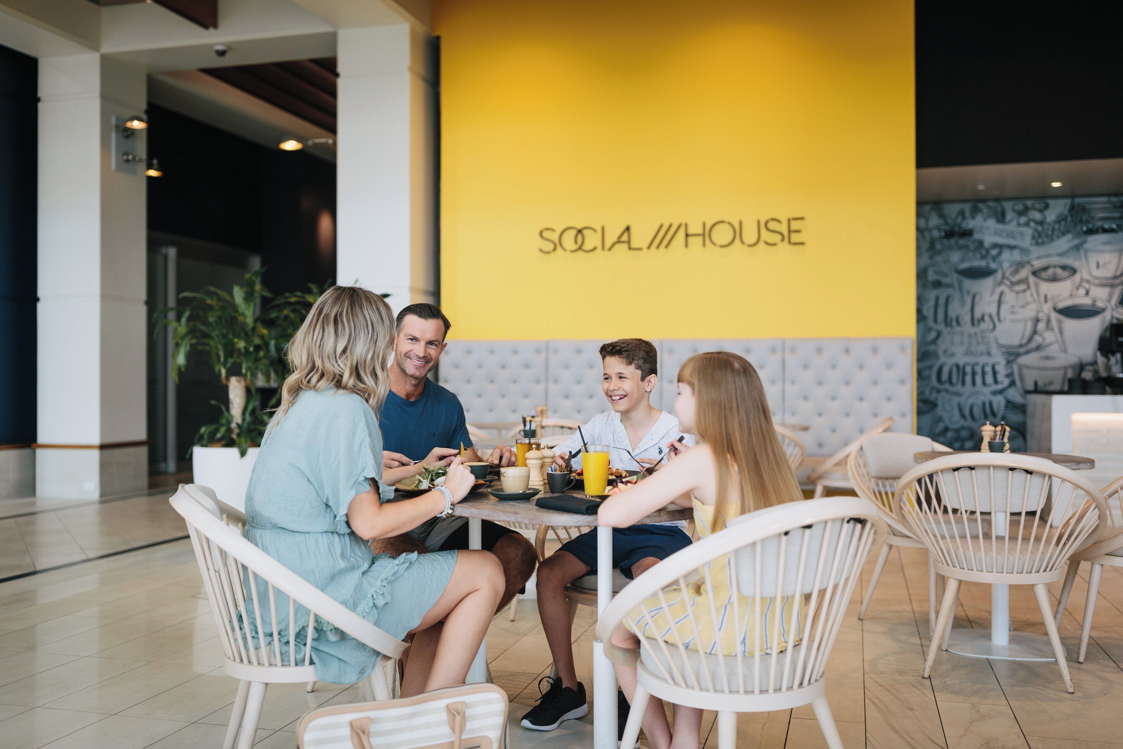 A group of people dining at a table with a yellow wall in the background labeled 'SOCIA//HOUSE'.
