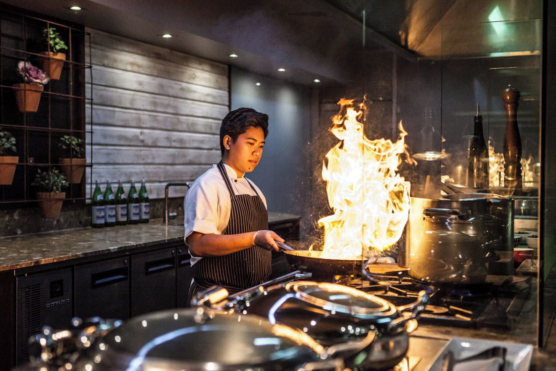 A chef performing a flambé with a pan on a stove in a kitchen.