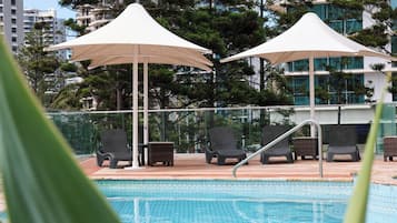 A swimming pool with lounge chairs and umbrellas, surrounded by tall buildings and trees.