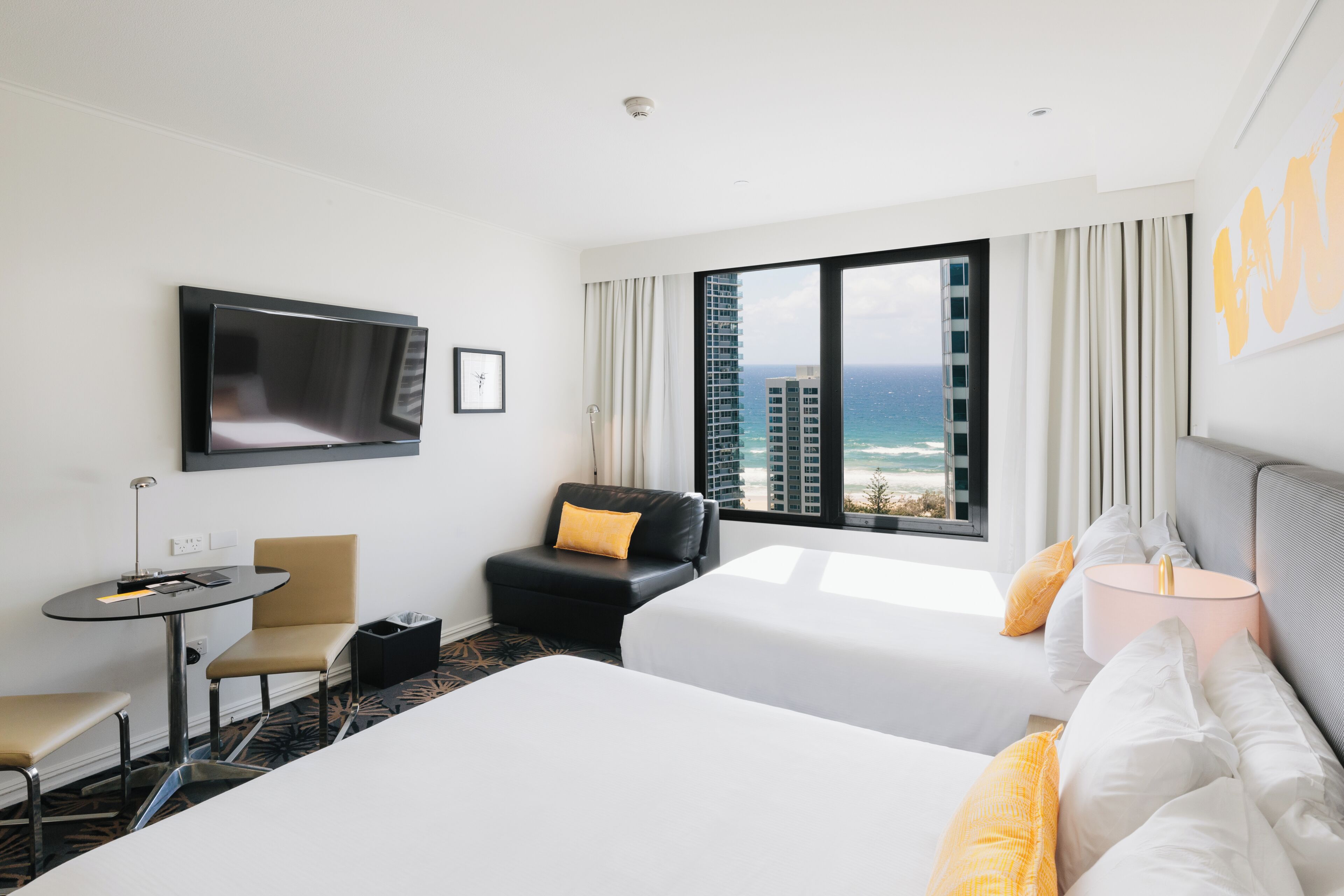 Ein Hotelzimmer mit einem Bett, einem Schreibtisch, einem Sessel, einem Fernseher und einem großen Fenster mit Blick auf das Meer und Gebäude.