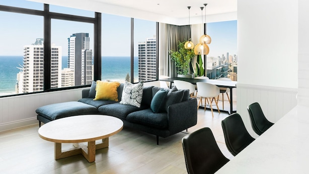 A modern living room with a large window, a sectional sofa, a round coffee table, and a dining area with chairs and a table. - Voco Gold Coast (Surfers Paradise)