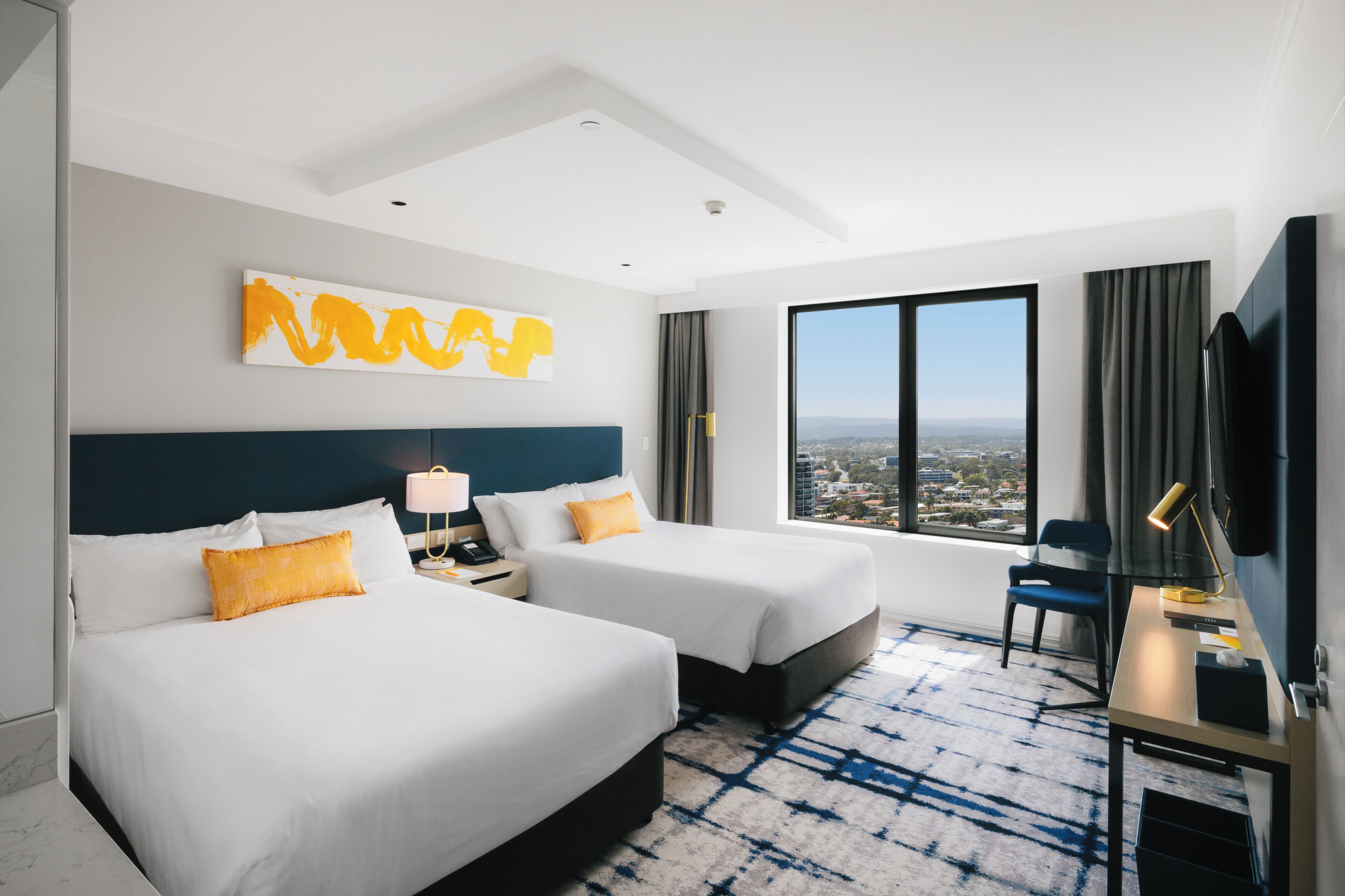 A hotel room with two beds, a desk, a chair, a TV, a large window with a city view, and a modern abstract painting on the wall.