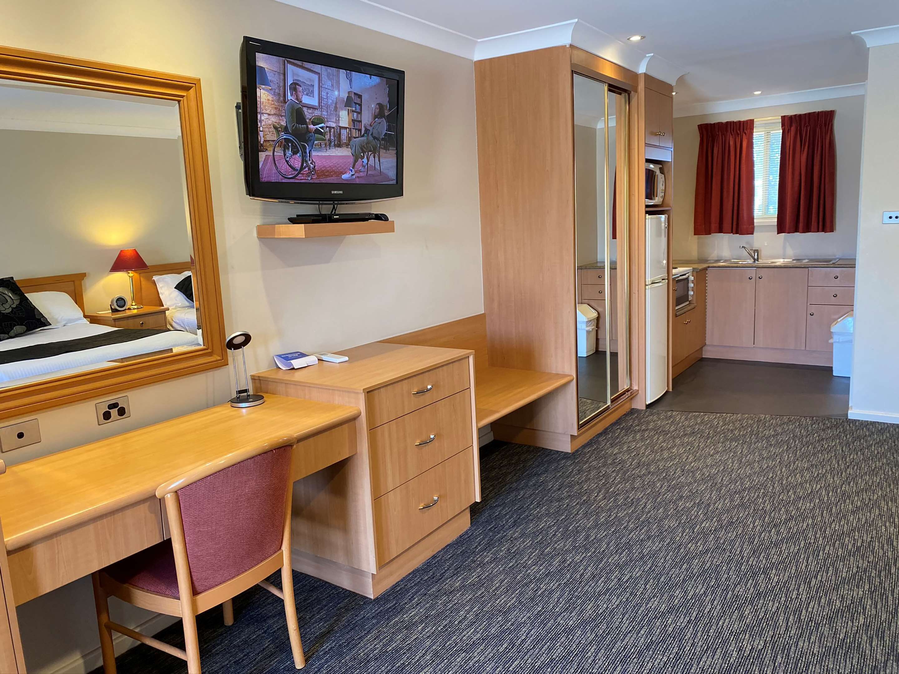 A hotel room with a bed, a desk, a chair, a TV, a wardrobe, a mirror, and a kitchenette.