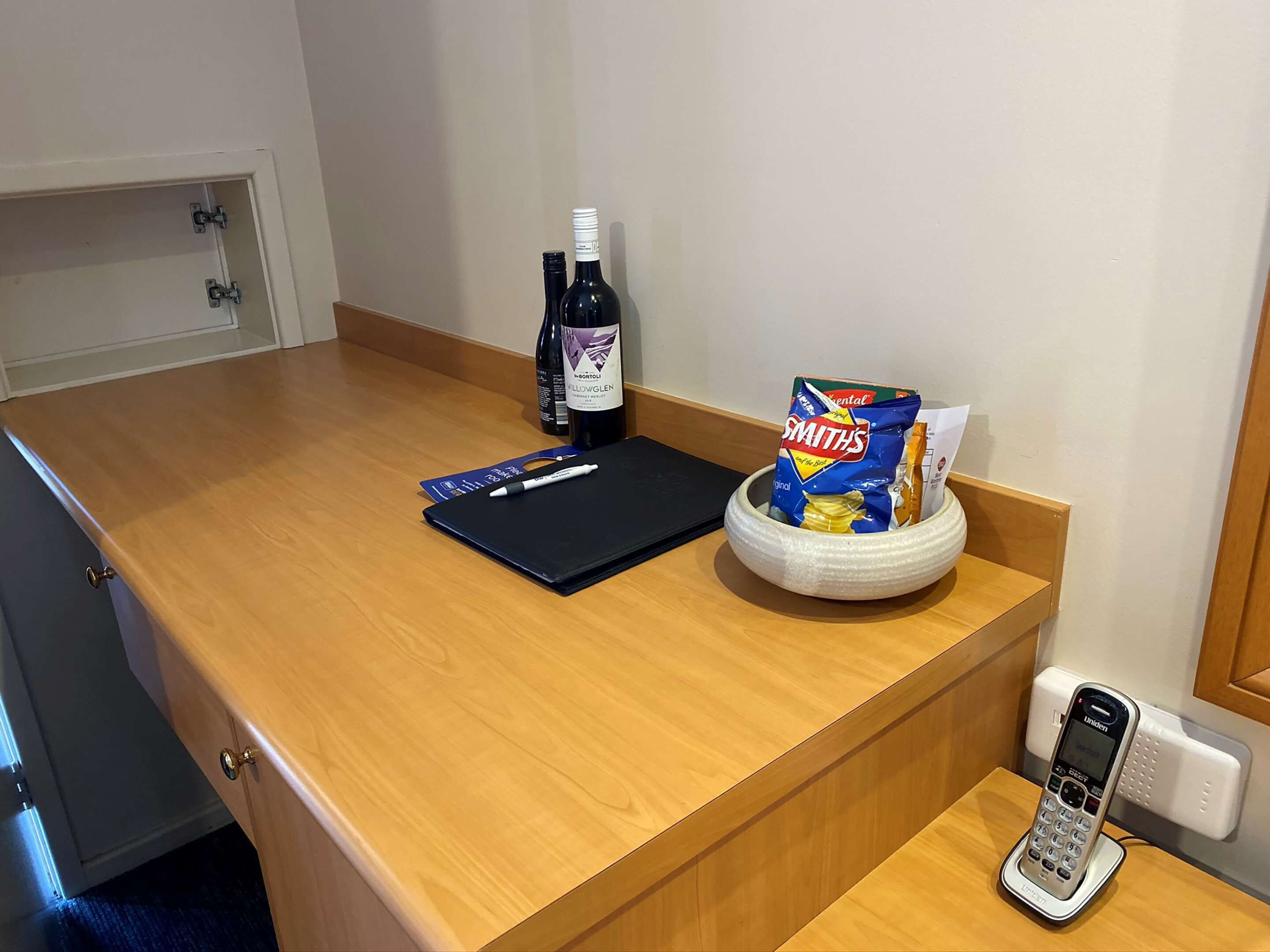 A wooden counter with a black notebook, a pen, a bottle of wine, a bag of chips, and a mobile phone.