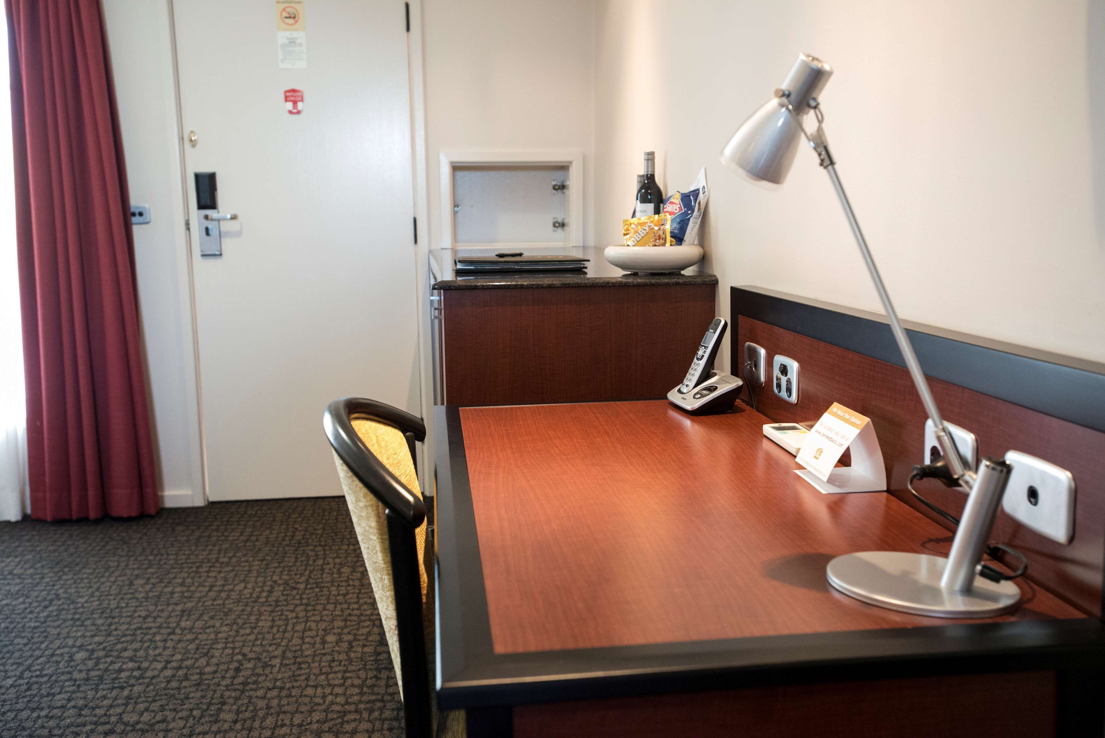 A hotel room with a wooden desk, a desk lamp, a chair, a small table with a bottle and snacks, and a television.