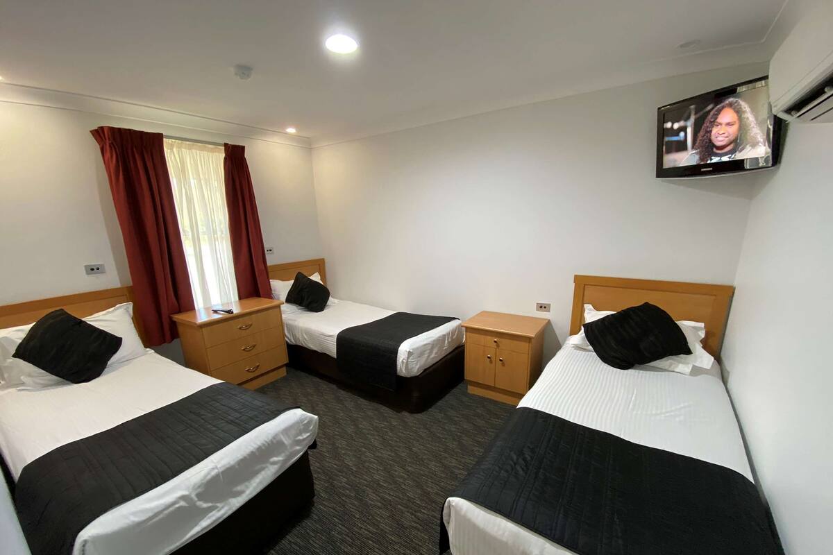 A hotel room with three single beds, a TV mounted on the wall, and a window with curtains.