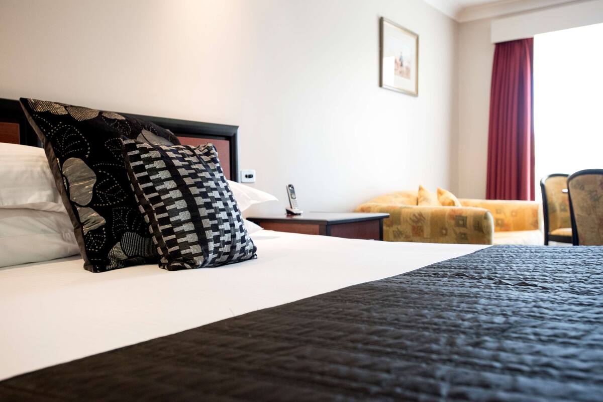 A hotel room with a bed, a nightstand, a lamp, a chair, and a window with curtains.