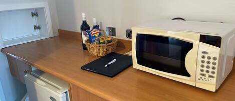 A kitchenette with a microwave, a basket containing snacks and a bottle, and a small refrigerator.