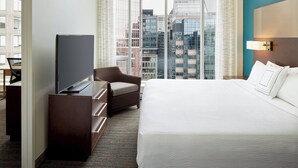 Habitación de hotel con una cama grande, un televisor sobre un aparador de madera, una silla y vistas de la ciudad a través de amplios ventanales.