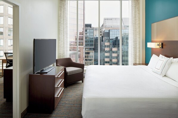 A hotel room with a large bed, a TV on a wooden dresser, a chair, and a view of the city through large windows. - Residence Inn by Marriott Montreal Downtown (Montreal)