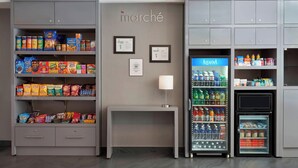 Una kitchenette con estantes llenos de bocadillos, un refrigerador y una mesita con una lámpara.