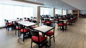 A spacious dining area with multiple tables and chairs, large windows, and a flat-screen TV mounted on the wall.