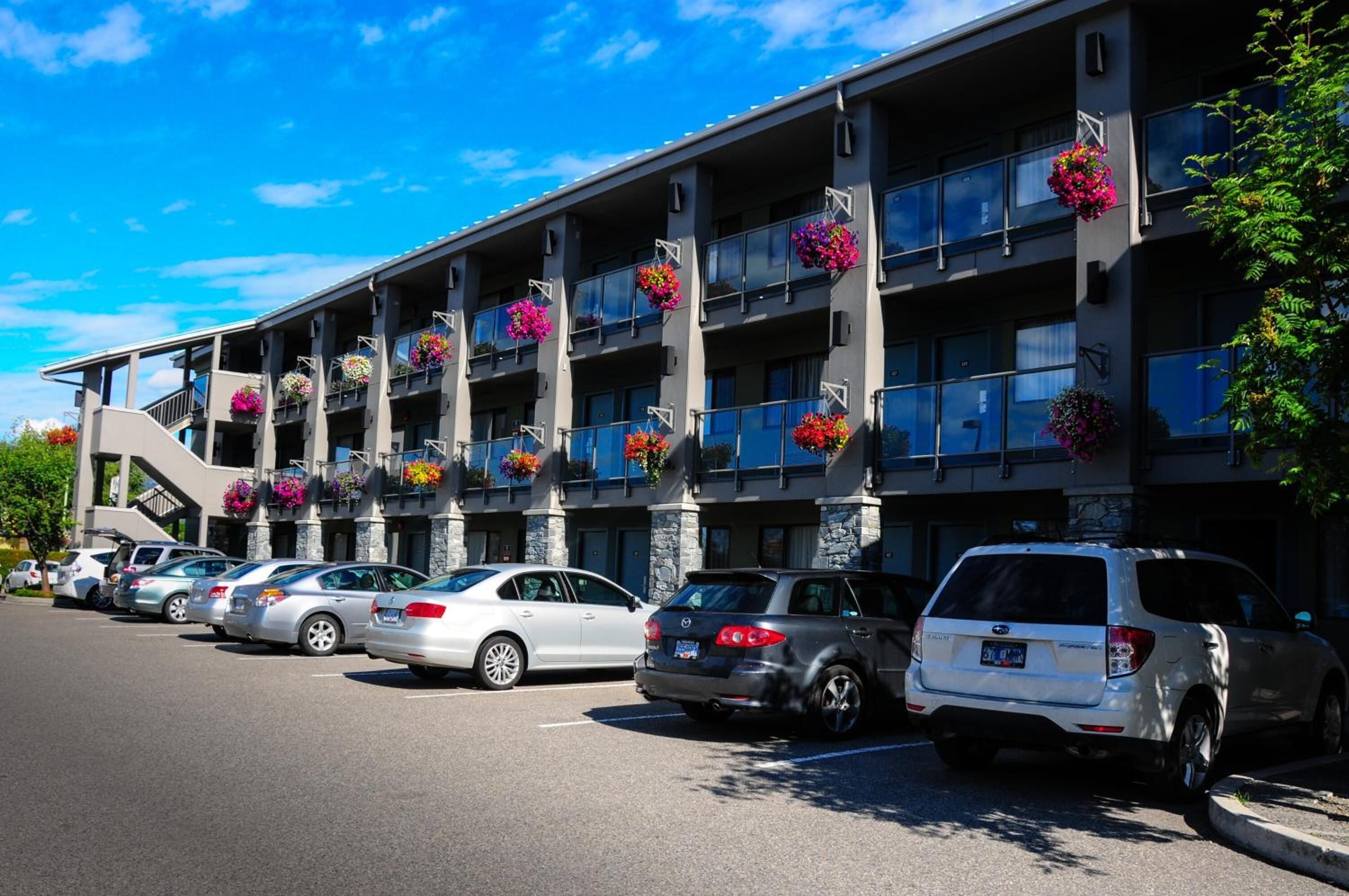 Un hotel de varios pisos con balcones decorados con macetas de flores, un estacionamiento con diversos vehículos y un cielo azul despejado.