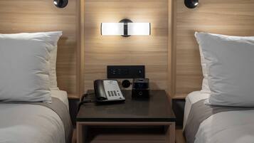 A hotel room with two beds, a bedside table with a phone and a digital clock, and a wall-mounted light fixture.