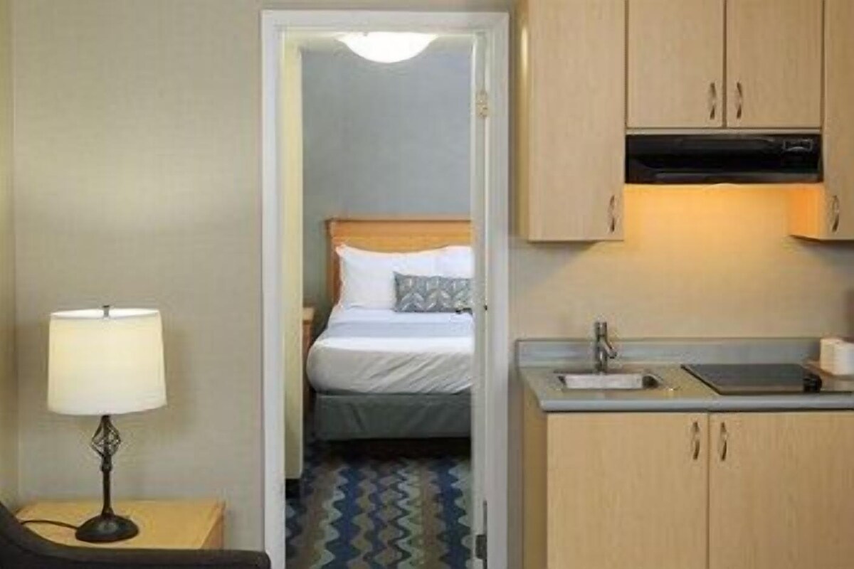 Habitación de hotel con cama, escritorio con lámpara, kitchenette con lavabo y puerta que da a un baño.