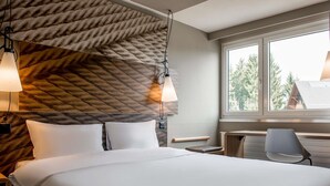 Habitación de hotel con cama, lámparas de noche, escritorio, una silla y ventana con vista a árboles.