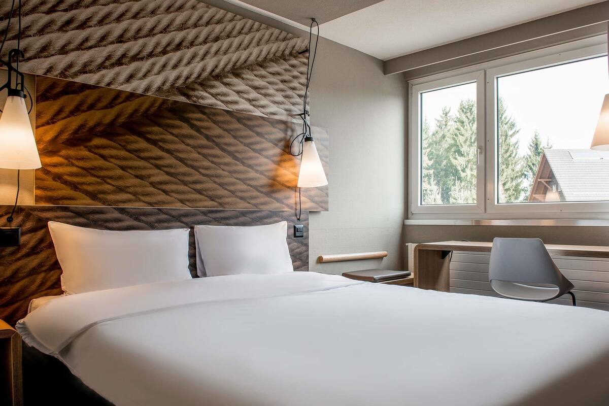 Ein Hotelzimmer mit Bett, Nachttischlampen, Schreibtisch, Stuhl und einem Fenster mit Blick auf Bäume.