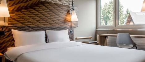 A hotel room with a bed, bedside lamps, a desk, a chair, and a window with a view of trees.